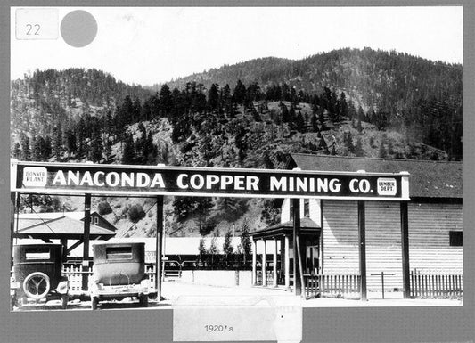 Black and white photo of Anaconda Copper Mining Co. lumber department entrance with vintage cars and forested mountain backdrop