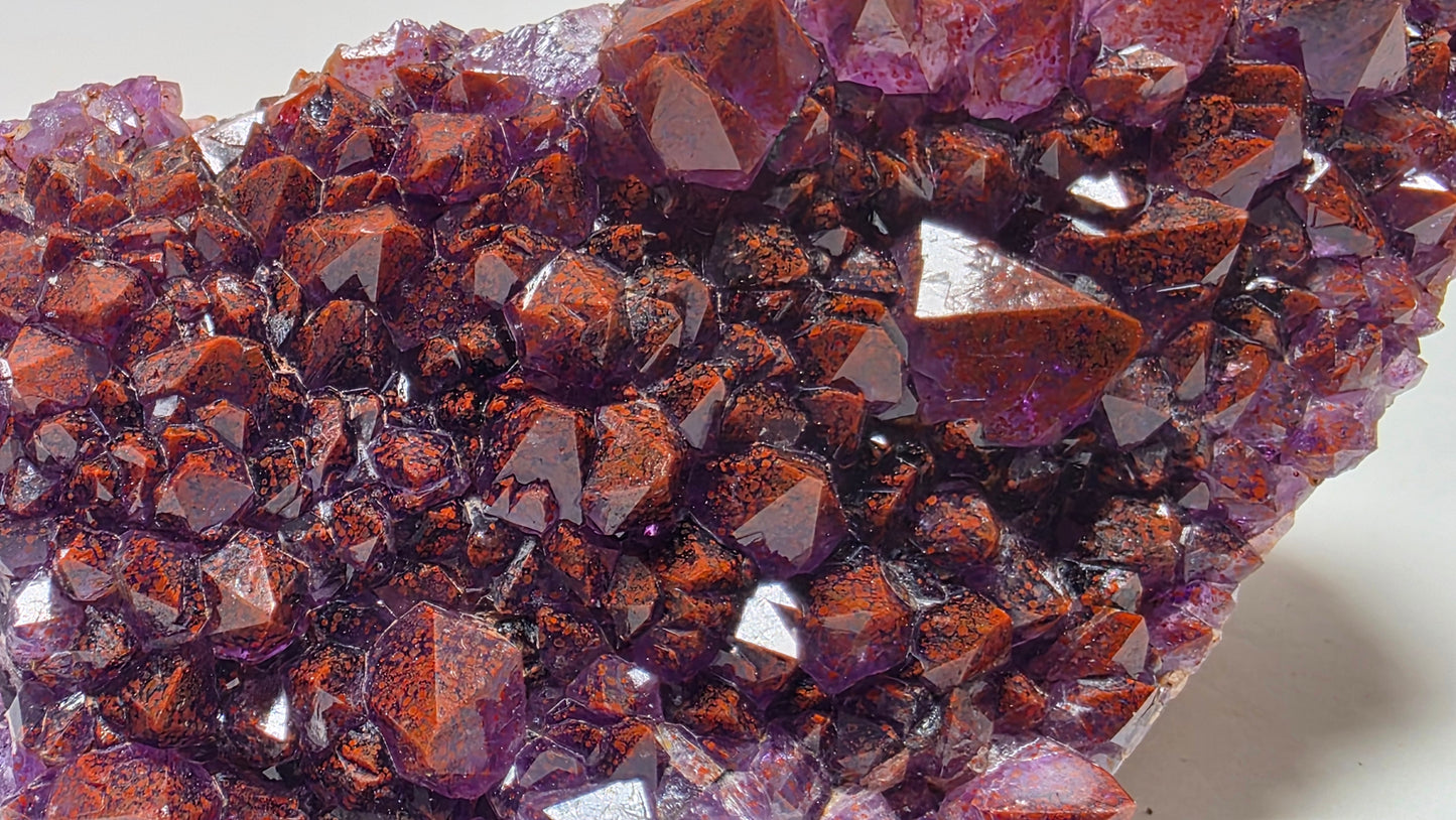 A close-up of a natural crystal cluster with faceted reddish-brown and purple mineral points, photographed on a plain light background.
