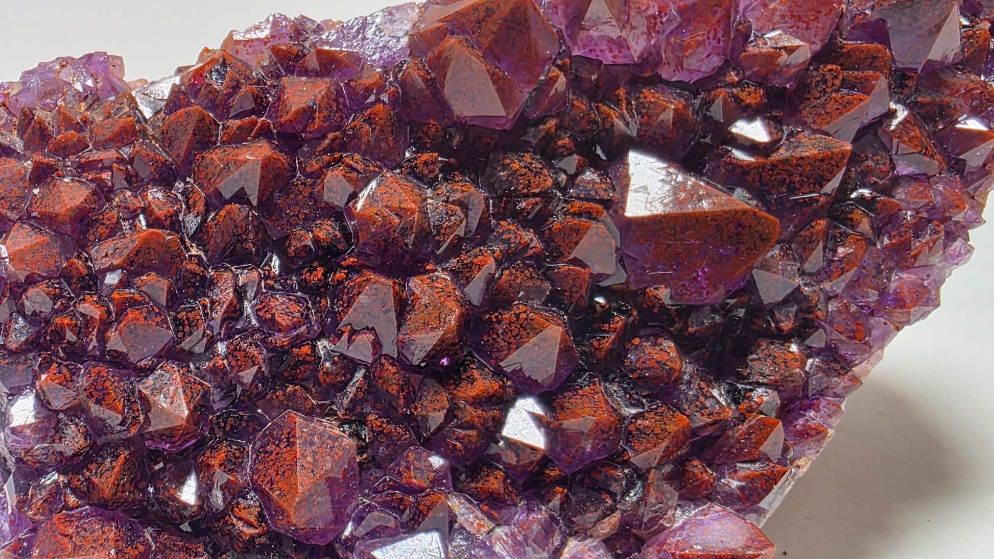 A close-up of a natural crystal cluster with faceted reddish-brown and purple mineral points, photographed on a plain light background.