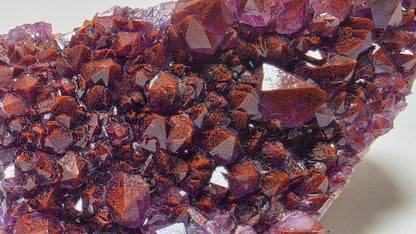 A close-up of a natural crystal cluster with faceted reddish-brown and purple mineral points, photographed on a plain light background.
