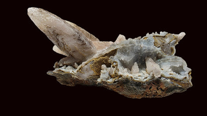 A natural mineral specimen featuring a large translucent pale crystal point rising from a rough, earthy matrix with gray and white crystalline areas, photographed on a plain light background.