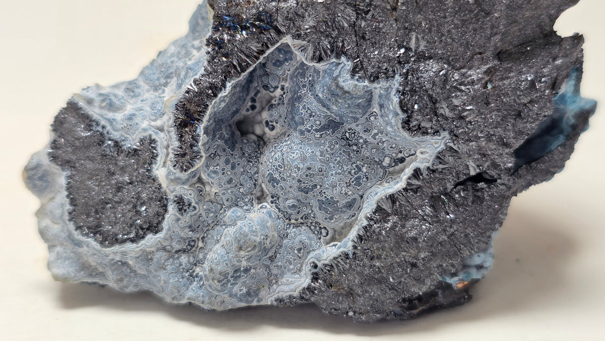 A close-up of a rough mineral or crystal specimen with a mottled mix of dark charcoal gray, pale icy blue, and white crystalline areas on a plain light background.