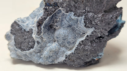 A close-up of a rough mineral or crystal specimen with a mottled mix of dark charcoal gray, pale icy blue, and white crystalline areas on a plain light background.