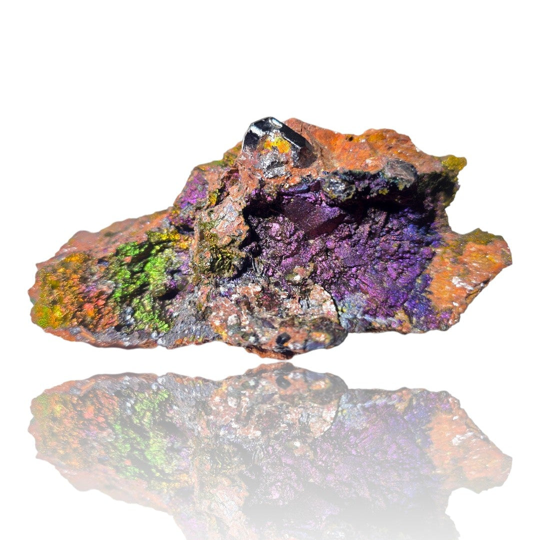 A raw, rough mineral specimen with a highly textured, porous surface, featuring vibrant metallic purple, orange, green, and yellow hues, displayed on a reflective white surface.