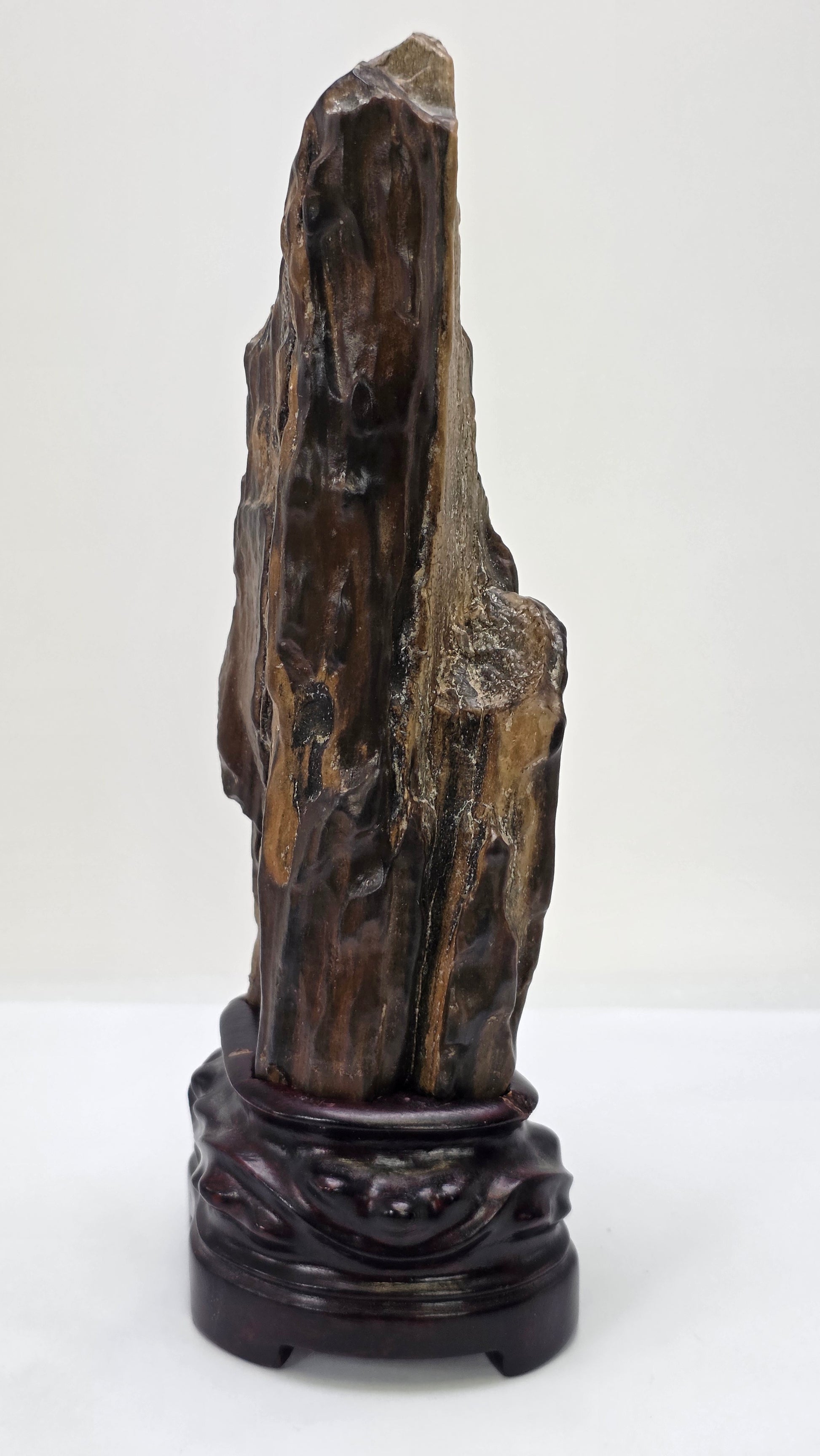 A vertical, dark brown and black petrified wood specimen mounted on an ornate, dark-stained carved wooden base. The stone features deep vertical ridges and a polished, glassy surface, tapering to a rugged point at the top. The entire piece stands 21 cm tall against a plain white background.