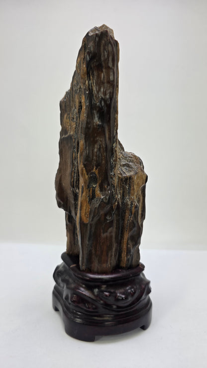 A vertical, dark brown and black petrified wood specimen mounted on an ornate, dark-stained carved wooden base. The stone features deep vertical ridges and a polished, glassy surface, tapering to a rugged point at the top. The entire piece stands 21 cm tall against a plain white background.