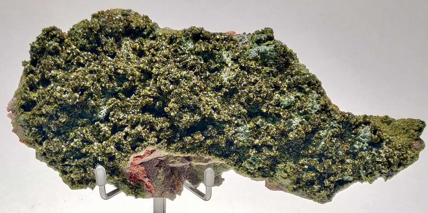 A deep olive-green Epidote crystal cluster with a vitreous luster, set against its natural matrix, from Hakkçari Province, Turkey, displayed on a metal stand.