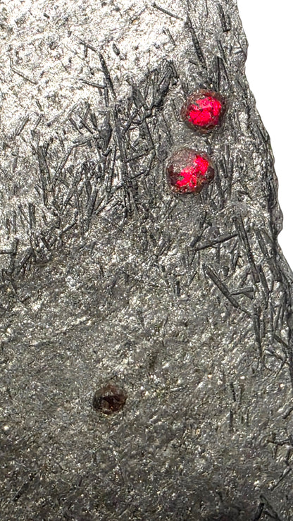 A close-up of a textured gray stone or mineral surface with dark needle-like inclusions and two small bright red gemstone accents near the upper right.