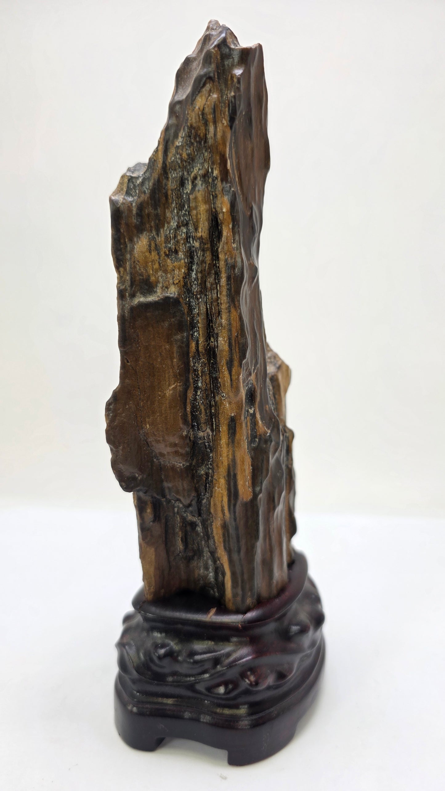 A vertical, dark brown and black petrified wood specimen mounted on an ornate, dark-stained carved wooden base. The stone features deep vertical ridges and a polished, glassy surface, tapering to a rugged point at the top. The entire piece stands 21 cm tall against a plain white background.